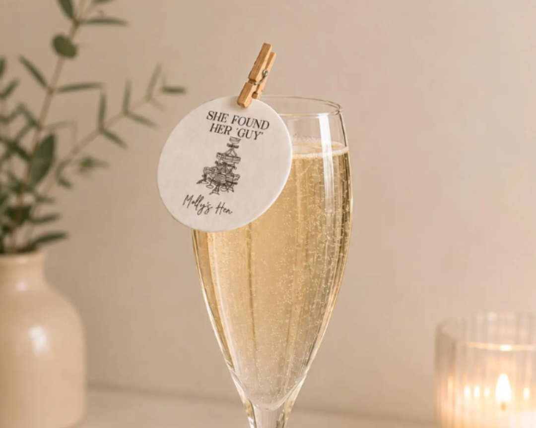 A clear champagne flute filled with bubbly champagne on a light marble surface, with a round white paper tag clipped to the rim. The tag has black text reading 'SHE FOUND HER GUY Happy's Mom' with a small tree illustration.